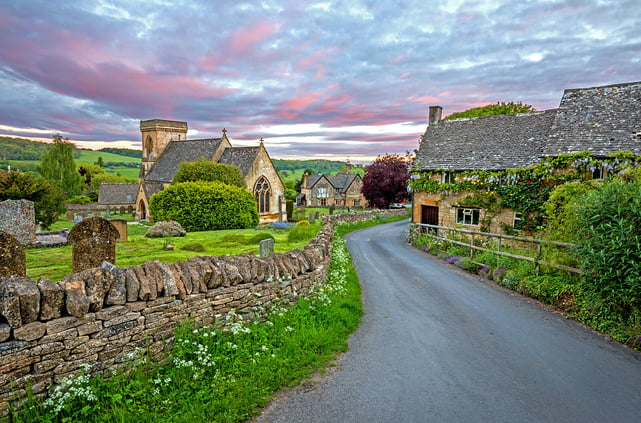Looking down an old village lane in the Cotswolds, England. An ancient stone church and graveyard stand on the left, with old stone cottages to the right.