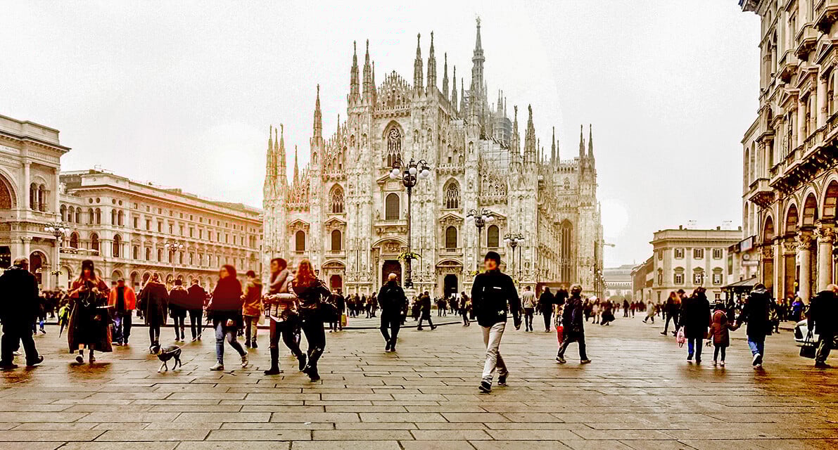 A view of the majestic, white, gothic style Duomo in Milan on a busy, cloudy day.
