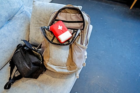 Image of a backpack on a sofa, top open, with a first aid kit resting on top. A smaller waist pack sits beside it.